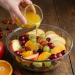 Pouring citrus dressing over Thanksgiving Fruit Salad with apples, grapes, pecans, and cranberries
